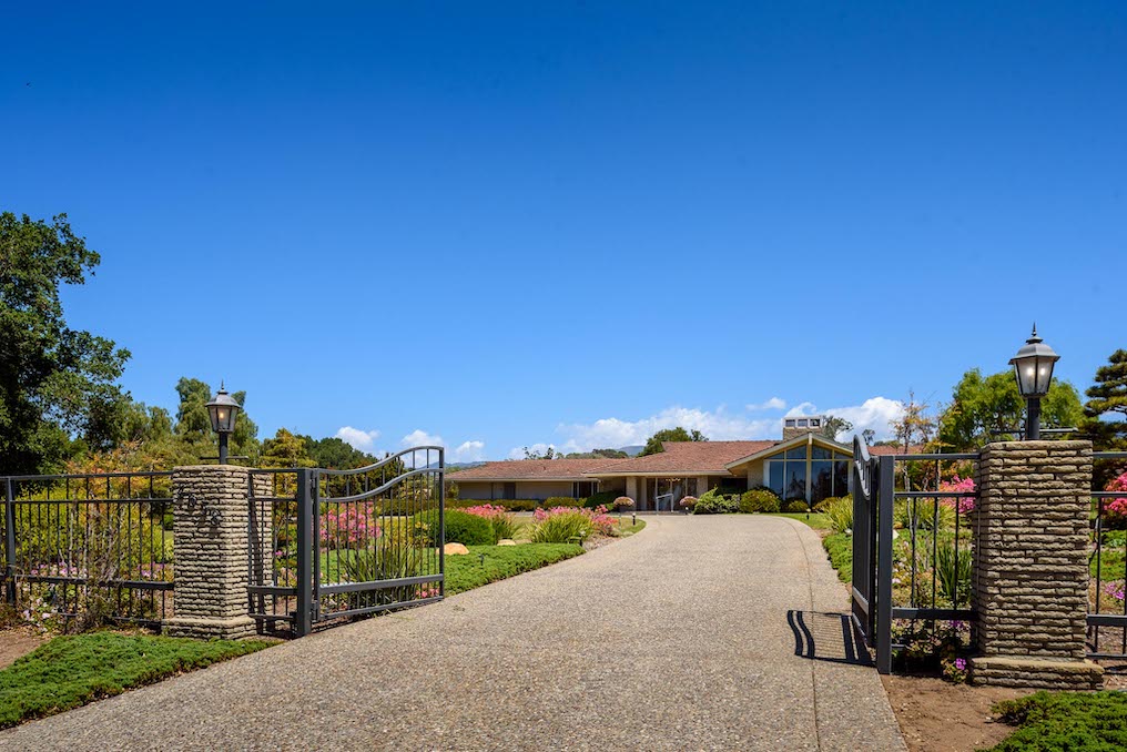 Beautiful iron gates the lead to a single story estate with lush landscaping and a winding driveway