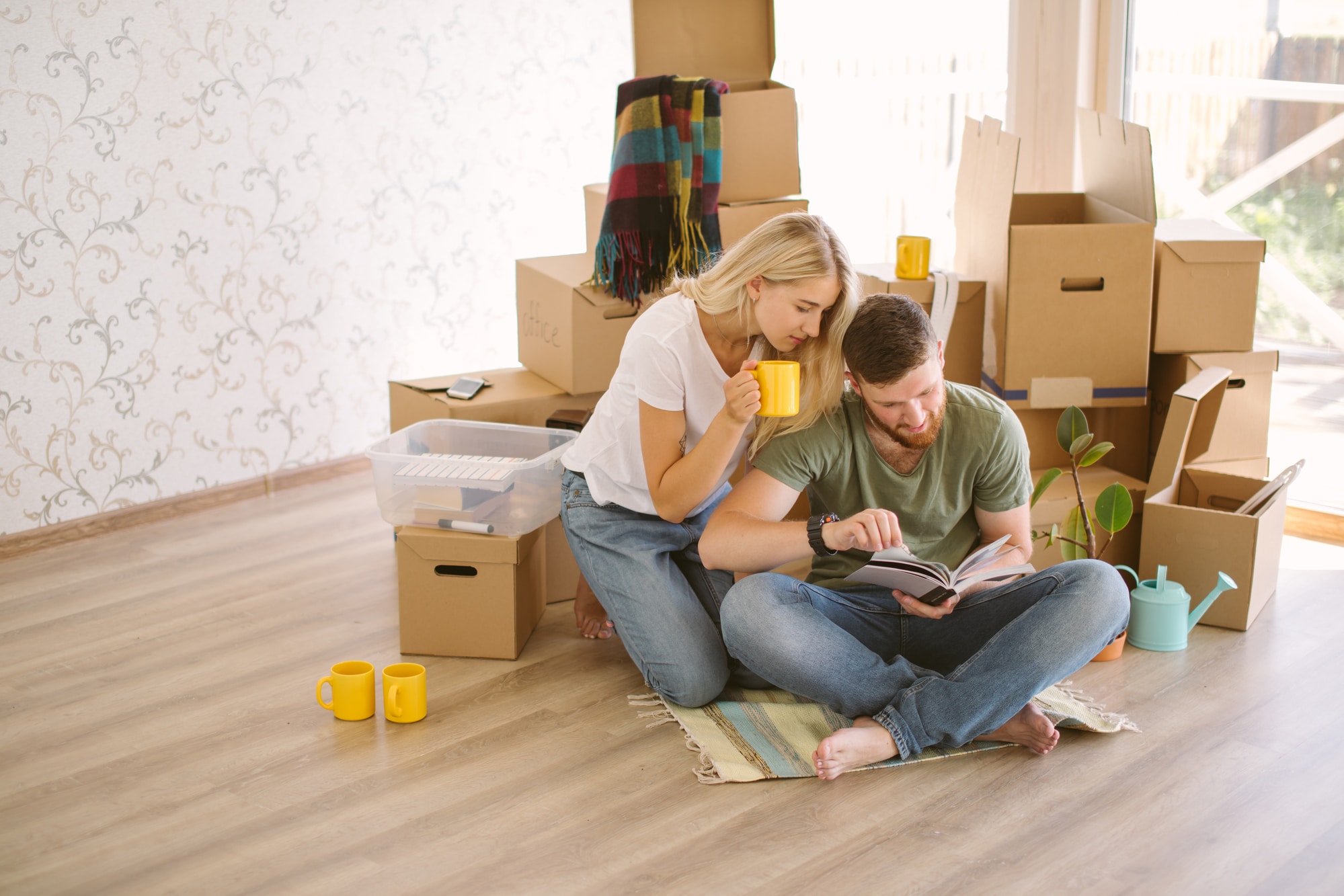 Young couple sitting on floor amid stacks of boxes, reading book about how to declutter your home