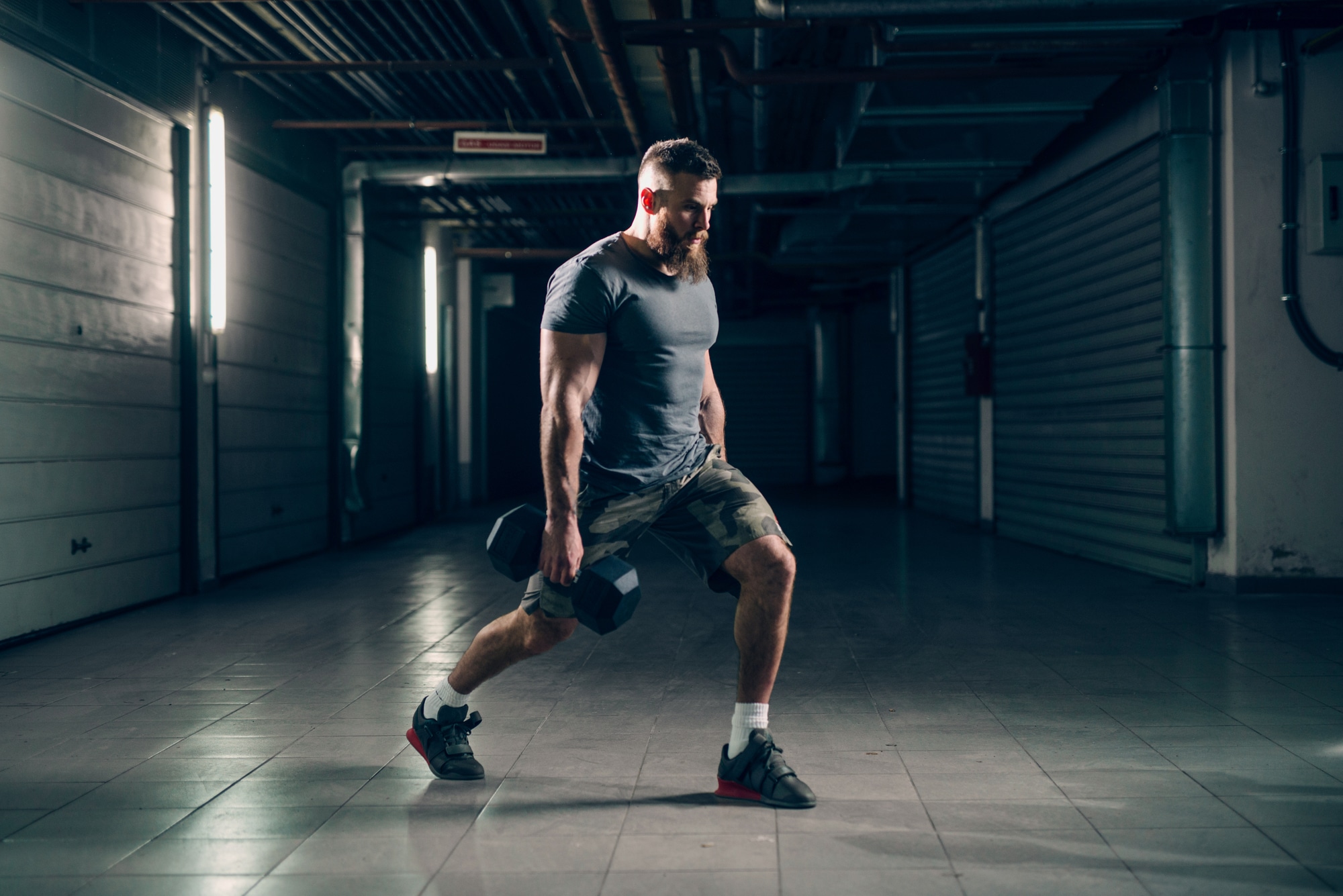 Side view of muscular attractive caucasian bearded man doing lunges with dumbbells in underground hallway. Storage units and pipes in background.