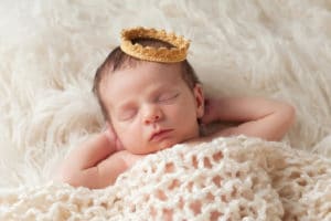 Portrait of a 12 day old newborn baby boy wearing a gold crown. He is sleeping on a beige flokati rug with his hands behind his head to illustrate Baby Checklist