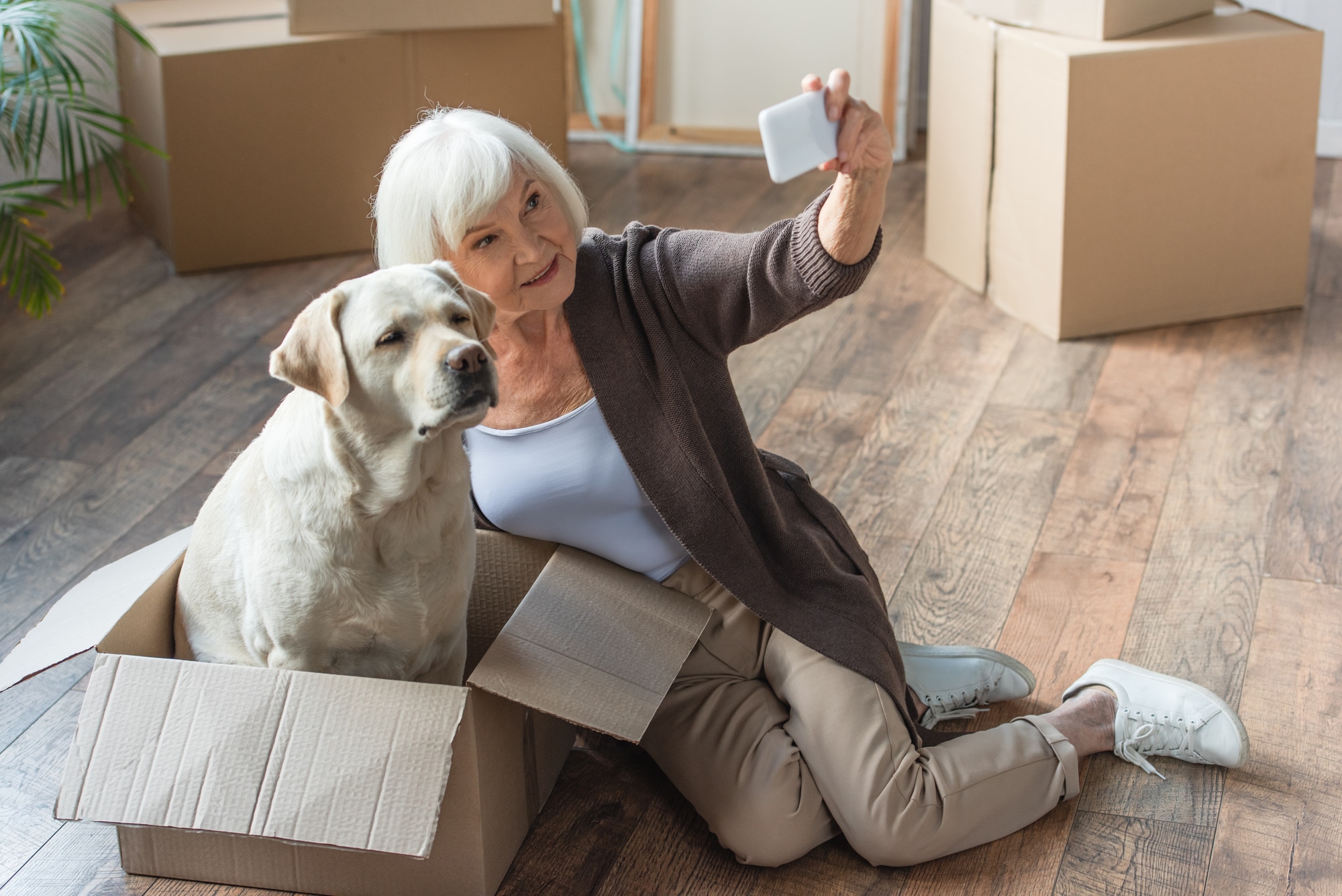 Older woman taking a selfie wit her dog who is sitting inside a moving box