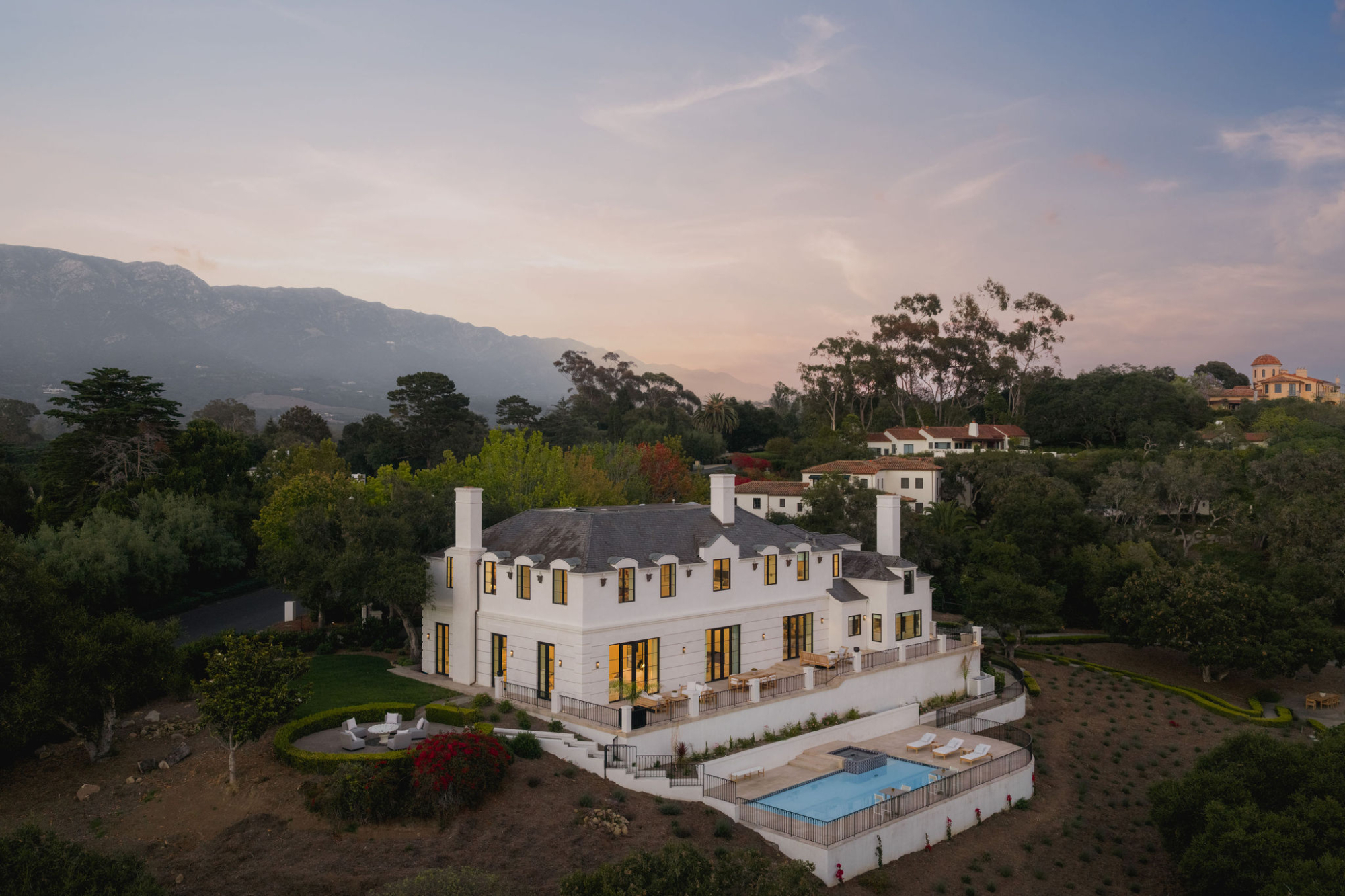 Twilight aerial view of a contemporary white Montecito estate with expansive terraces, a swimming pool, and landscaped grounds, set against the Santa Ynez Mountains beneath a colorful evening sky.