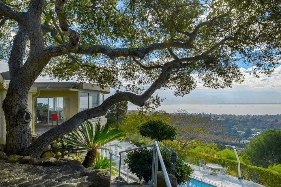 A section of a mid-century home, with floor-to-ceiling windows, a large oak tree in the fireground and the ocean in the background.
