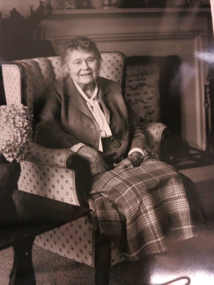 A black and white photo of architecut Harriet Moody. taken during her older years, sitting in front of a fireplace with a plaid blanket over her lap.