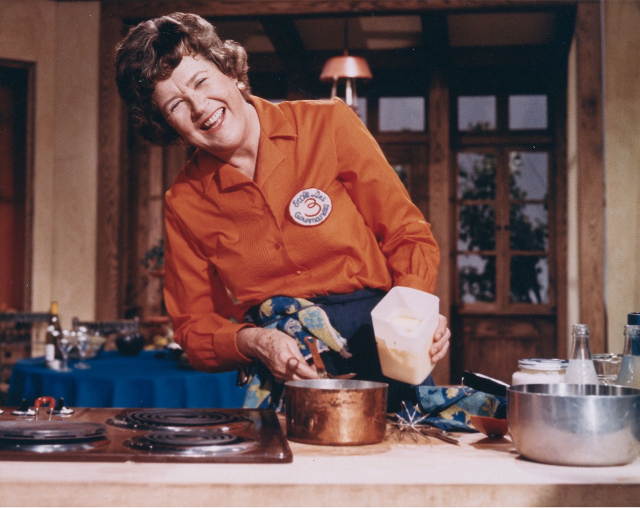 Julia Child in the kitchen cooking, wearing a bright orange shirt and a big smile, holding a copper saucepan.