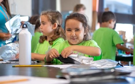 Two young girls sitting at a table in green t-shirts doing an art project.