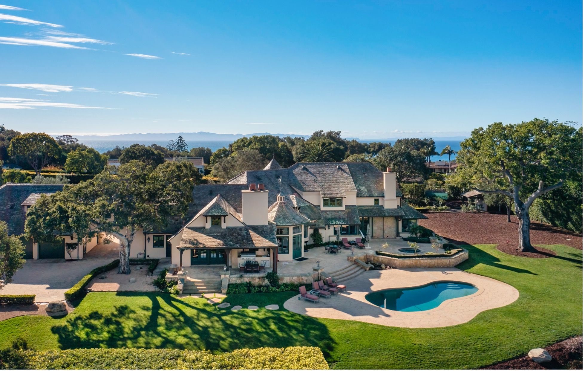 A French Country estate in Hope Ranch, with a kidney shaped pool surrounded by green grass and beautiful landscaping in the foreground and the large home in the background and blue sky above