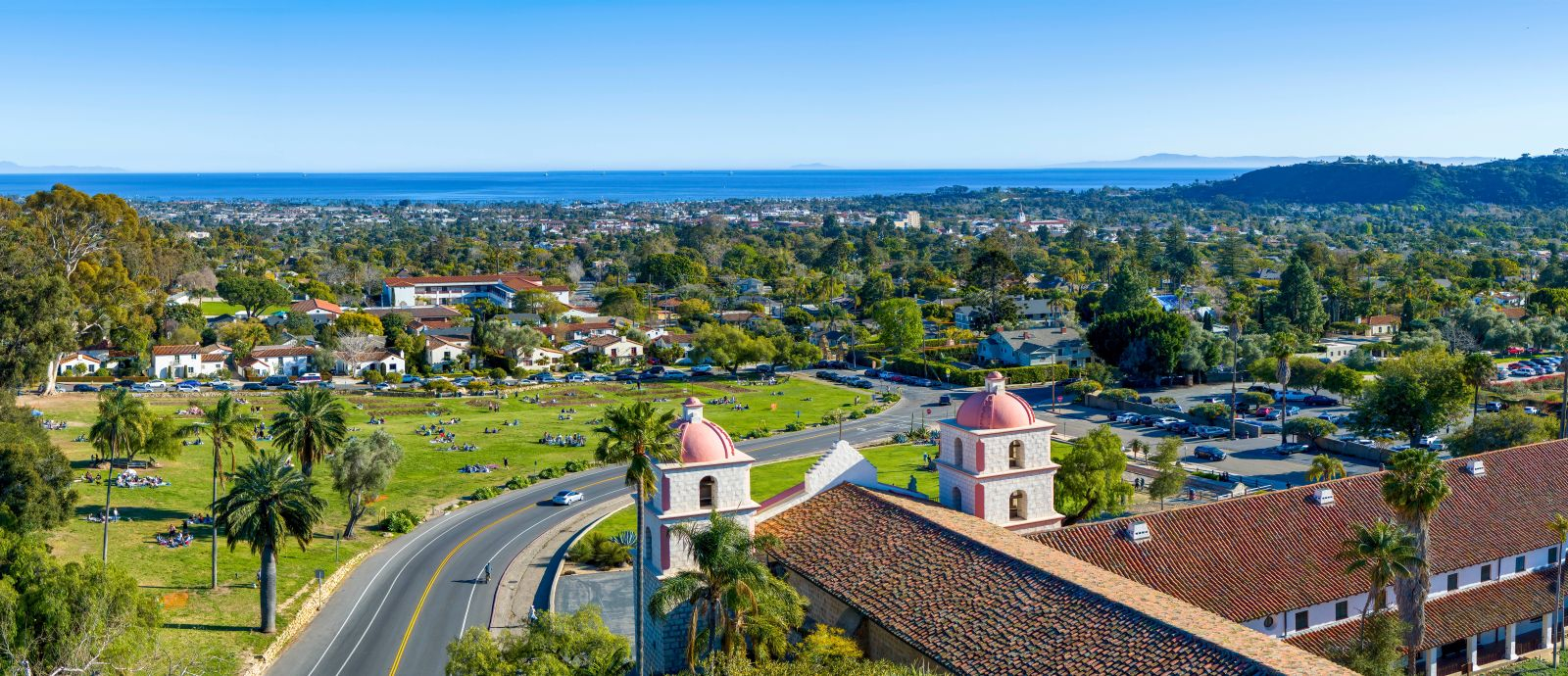 Aerial view of the historic Santa Barbara Mission with its iconic twin bell towers, palm trees, Mission Rose Garden, and sweeping views of downtown Santa Barbara, the Pacific Ocean, and the Channel Islands in the distance.