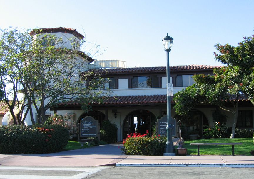 View of the facade of the Santa Barbara Airport Terminal with its white stucco walls and red tile roof, and a lawn and trees in front