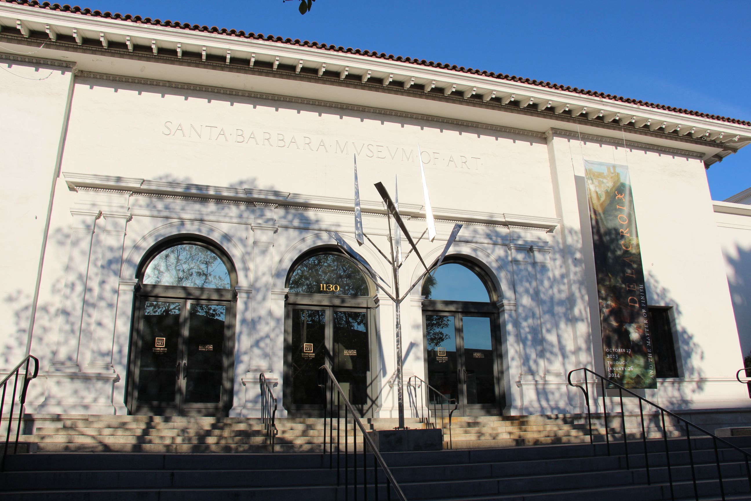 A portion of he facade of the Santa Barbara Museum of Art with a hint of its red tile roof, and blue sky.