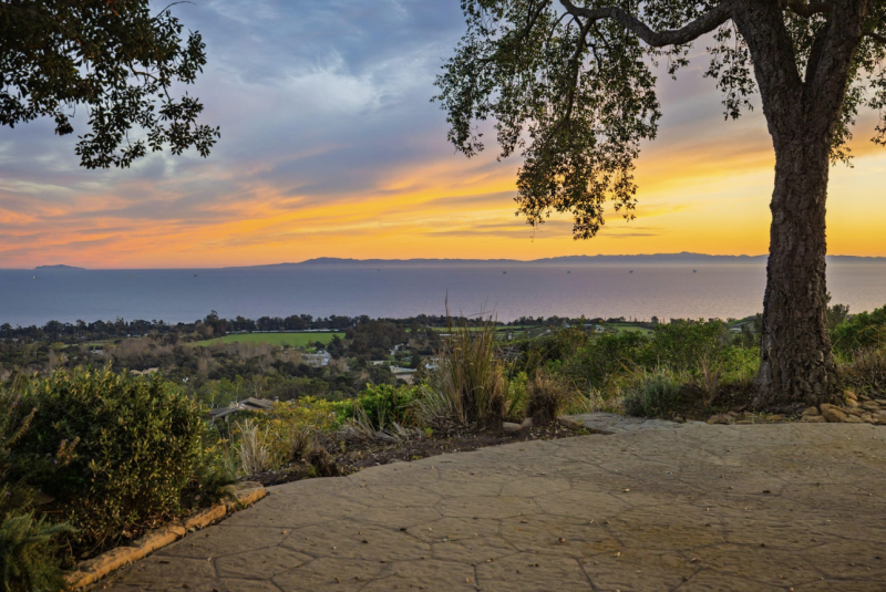 Panoramic ocean-view patio framed by mature oak trees overlooking the Santa Barbara coastline at sunset, with rolling green hills, colorful clouds, and the Pacific Ocean stretching to the Channel Islands.