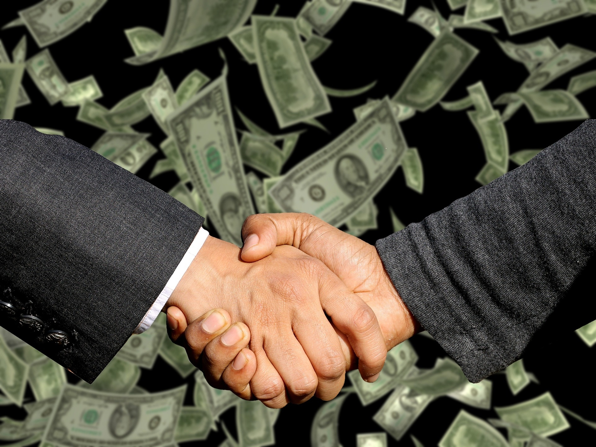 Forearms and hands of suited men shaking hands in front of falling currency.