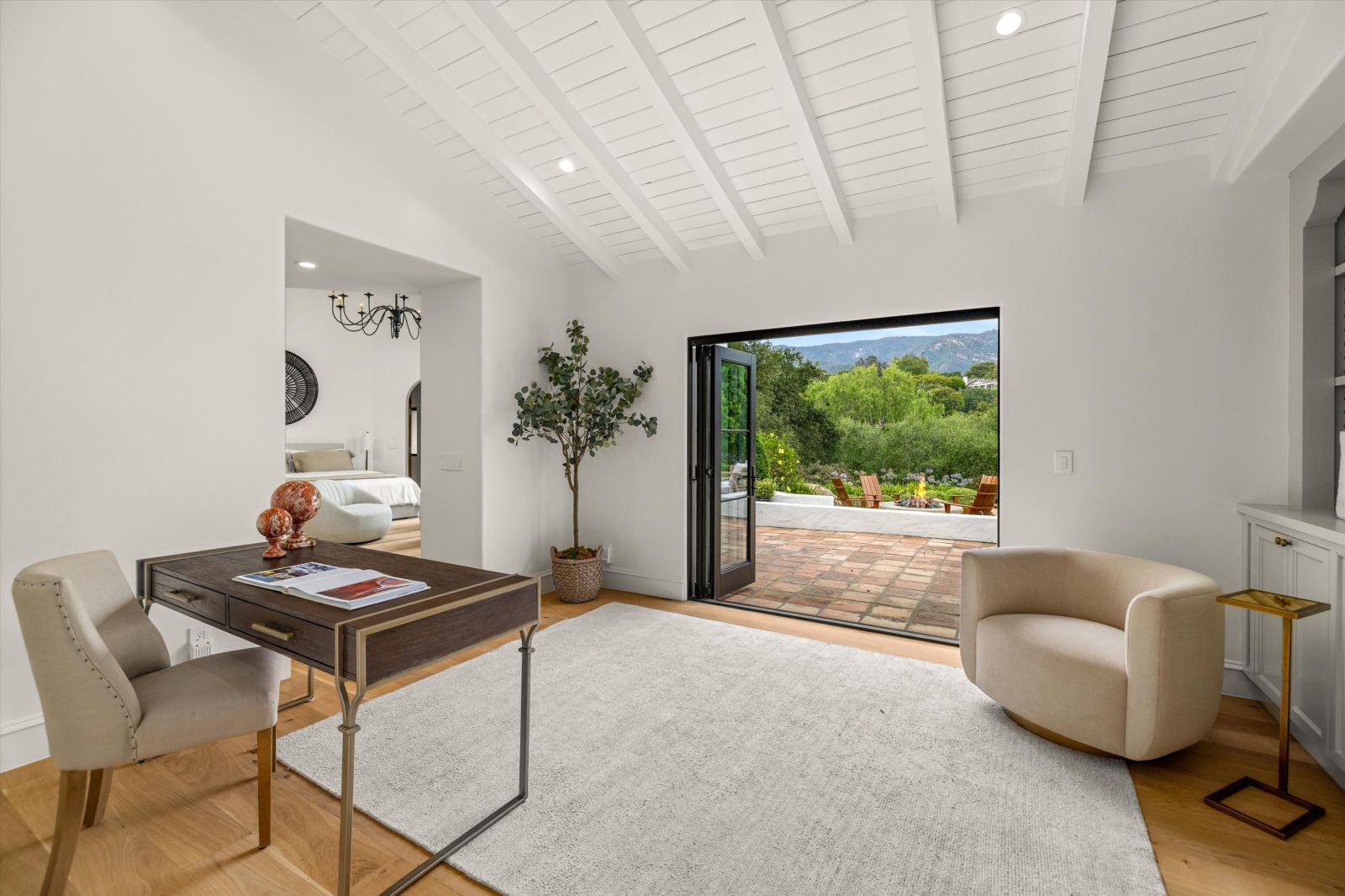 A white room with a vaulted ceiling, desk and a chair looking out past open doors to a terrace.