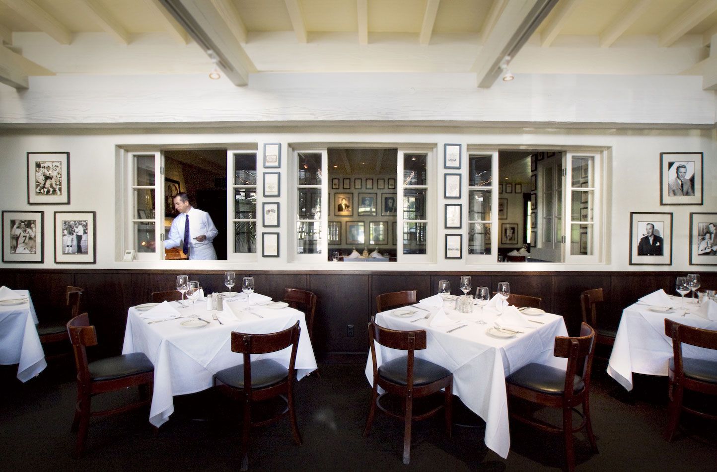 Looking in to the dining room at Lucky's Restaurant, with tables set with white linens and windows to the kitchen with a waiter prepping