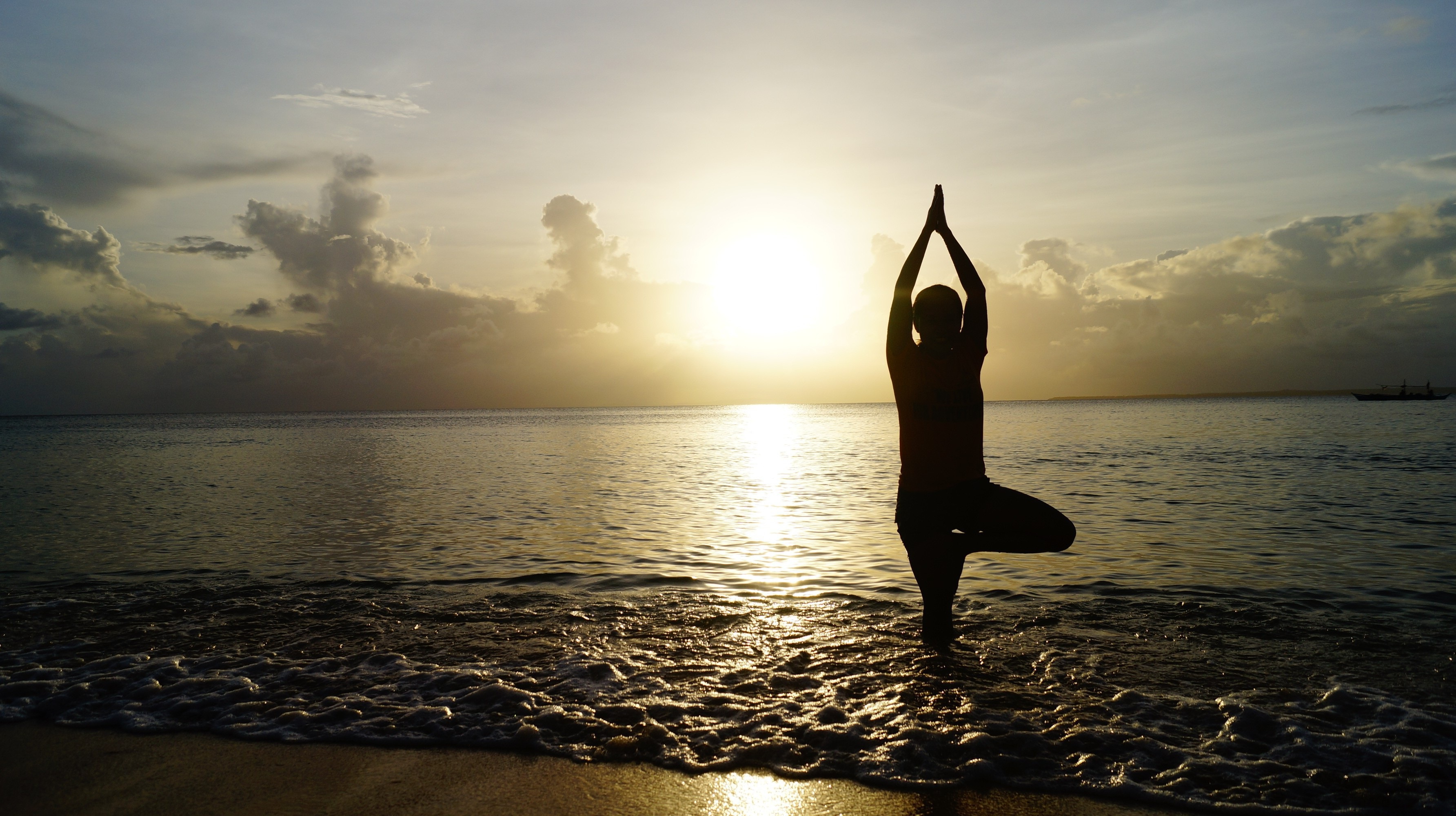 Silhouette of a person practicing tree pose while standing in the shallow ocean at sunrise, with gentle waves, glowing sunlight reflecting across the water, and dramatic clouds creating a peaceful, meditative scene.