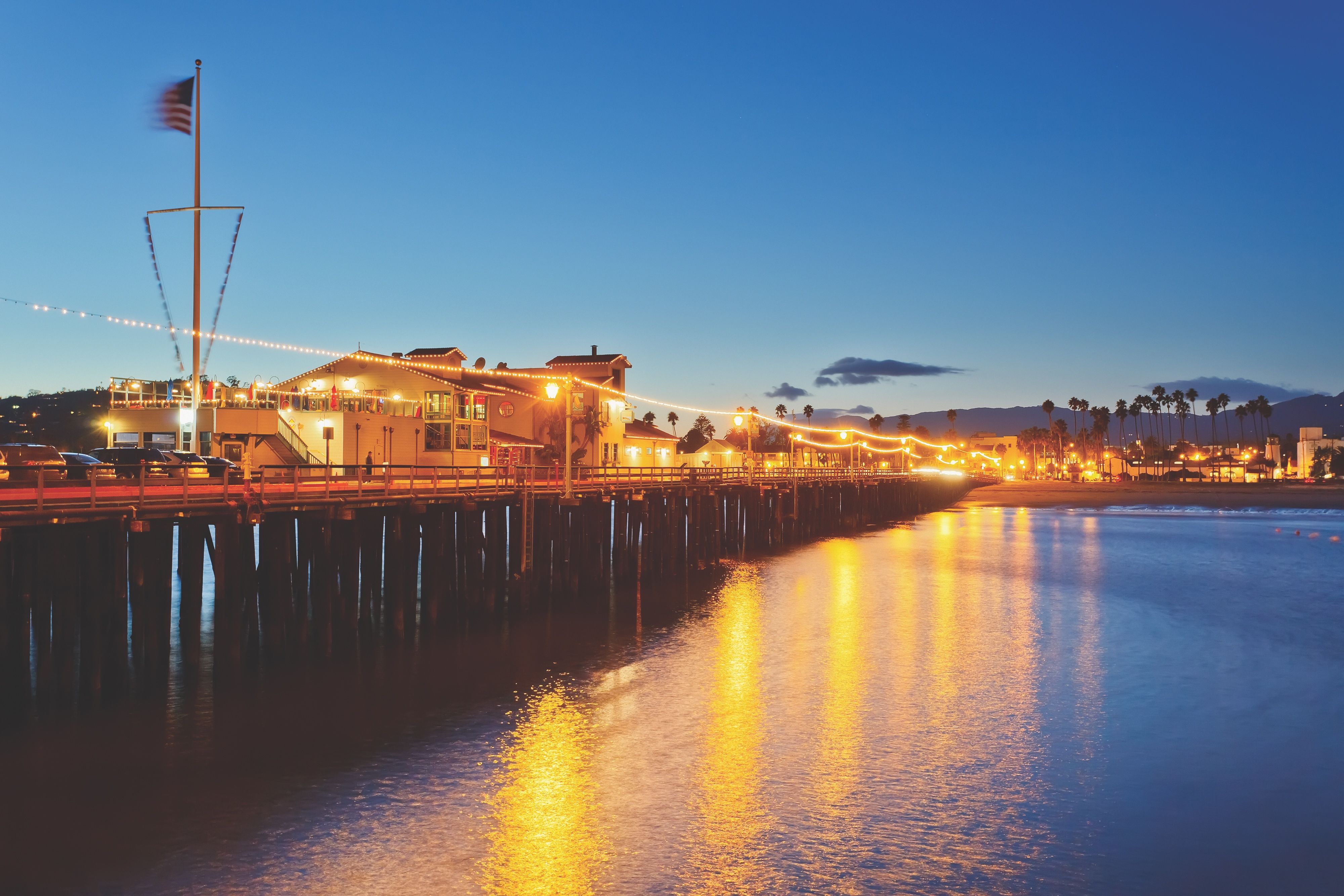 Santa Barbara Stearn's Wharf from the ocean end at dusk, with the wharf buildings illuminated and the ocean water spsarkling blue and gold.