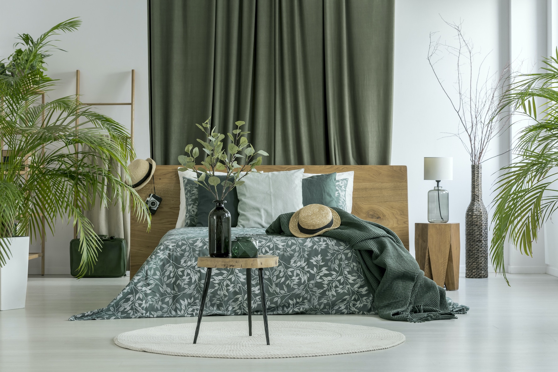 Table with plant on white round carpet and green curtain behind king-size bed with hat in bedroom with plants