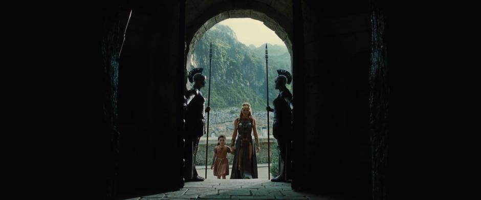 Hippolyta walks up the stairs of the tower holding young Diana's hand as they pass two guards.