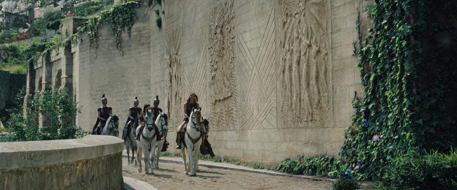 Hippolyta and young Diana ride down the street to the tower followed by several guards.