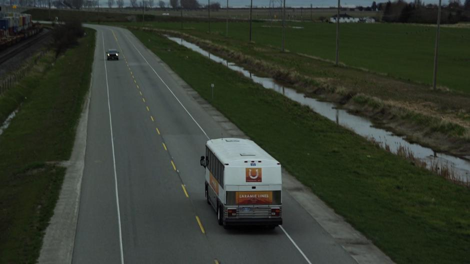 The bus drives away from the camera while a car passes the other direction and a train drives along the tracks to the left.