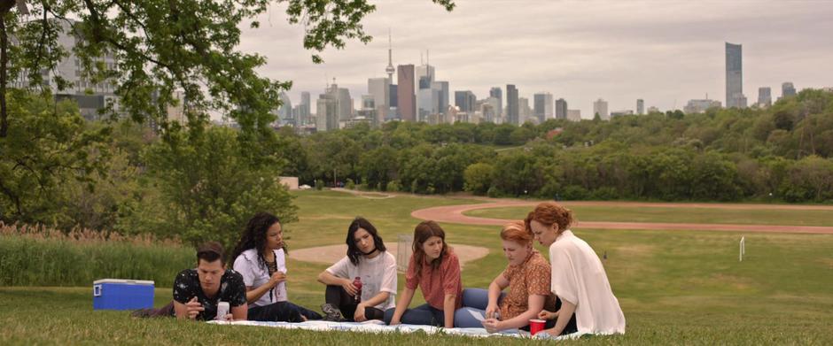 Kirsch, Mel, Carmilla, Laura, Lafontaine, and Perry sit on a blanked in the park while Laf and Perry examine a picture of the mansion.