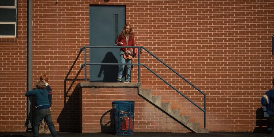 Max drops something in the trash can while standing atop some steps outside the building.