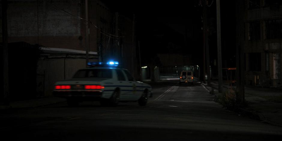 One of the police cars turns the corner and follows the van down the street towards the tunnel.