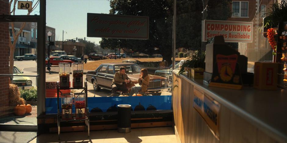 Bob and Joyce sit on the bench in front of the store eating their lunch.