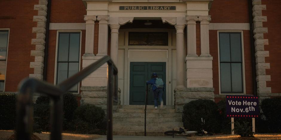 Dustin runs up the stairs to the front door of the library after dumping his bike on the ground.