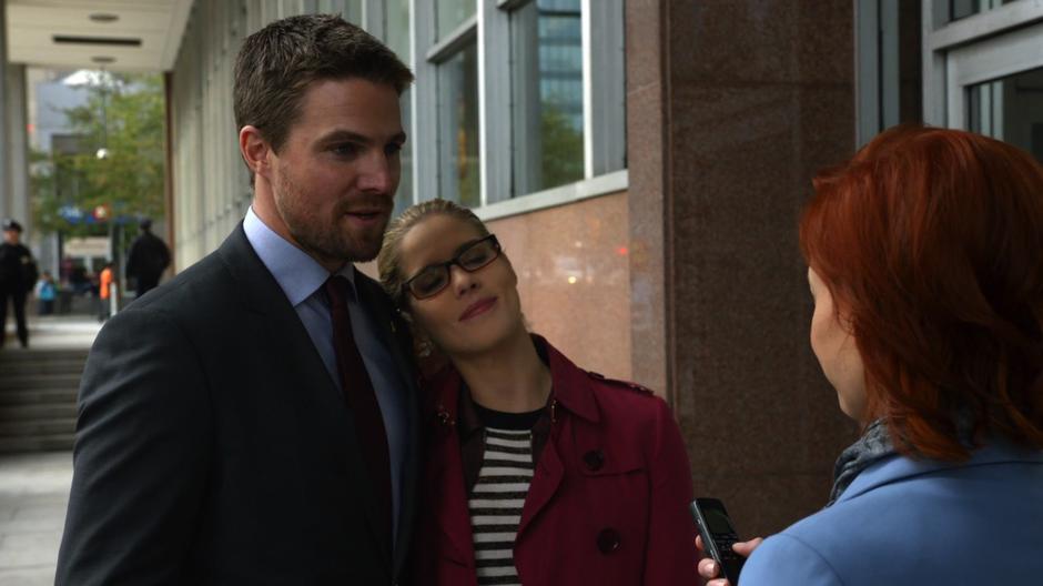 Felicity leans on Oliver's shoulder while he talks to the reporter.