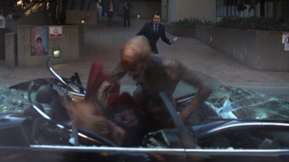 Kara fights with a dominator on top of a crushed car while the businessman watches.