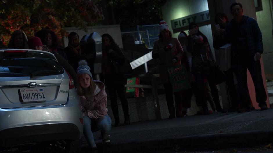 A young girl crouches behind a car as other bystanders watch from behind her.