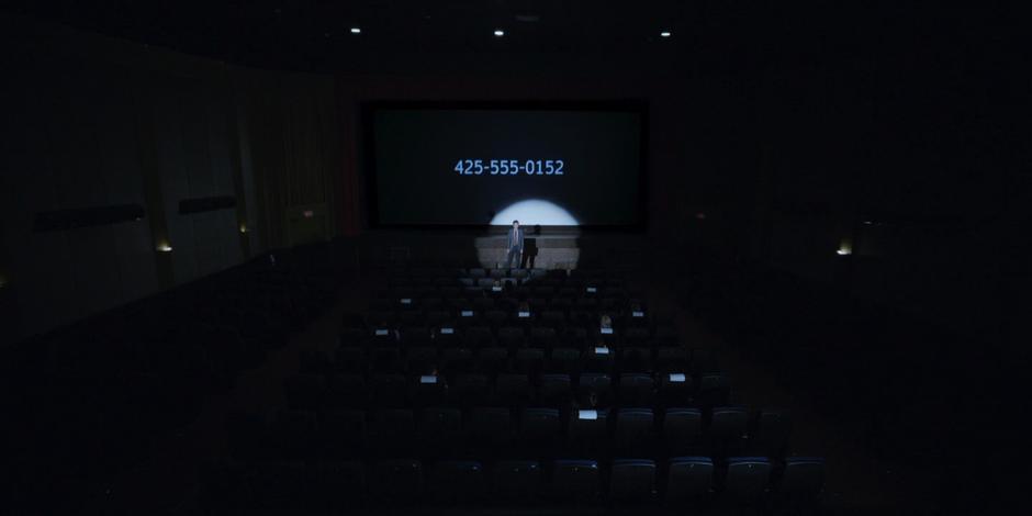 A man stands at the front of the theater preparing everyone for the update while a phone number flashes on the screen.