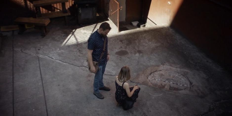 Marcy kneels next to the artwork on the ground and asks David about its artist.