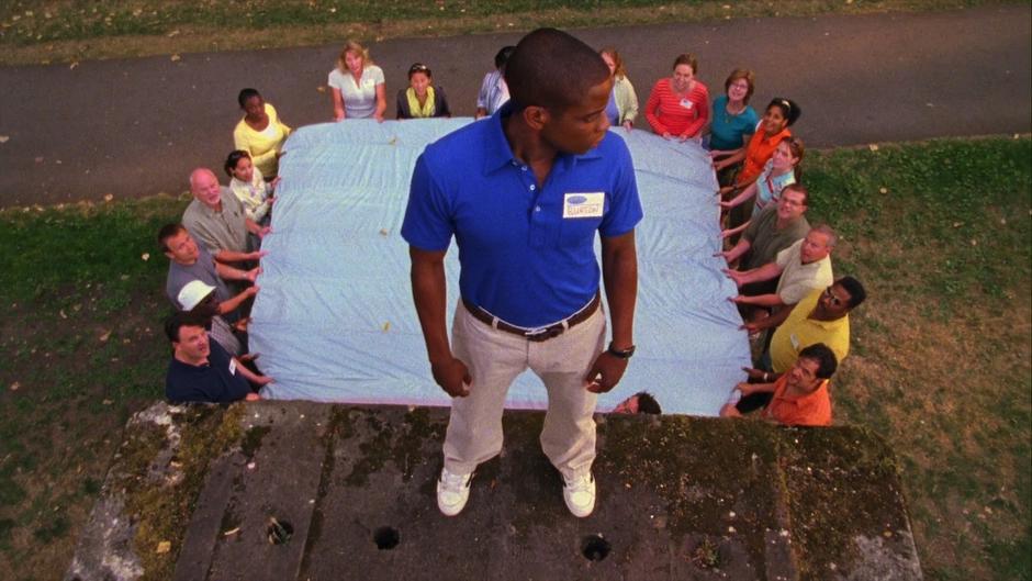 Gus stands atop a platform while his coworkers hold out a sheet behind him for the trust fall.