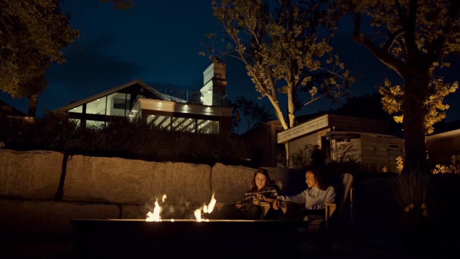 Jess and Heather sit side-by-side in front of the fire in the back yard in the evening.