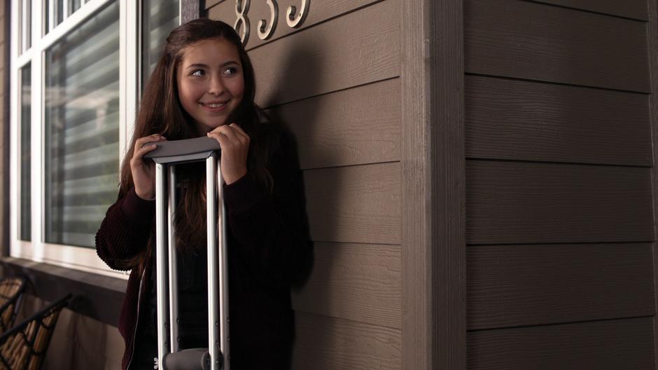 Ruby smiles while holding Alex's crutches out of sight of the door.