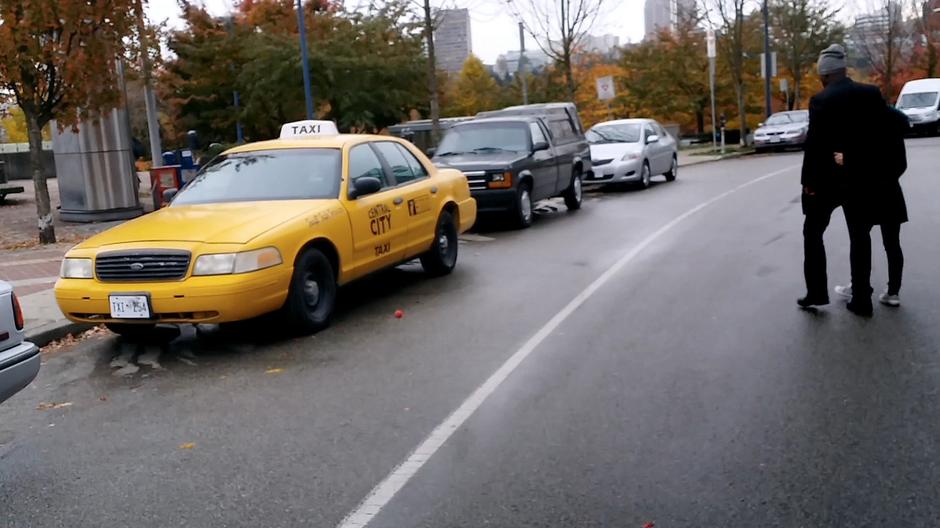 Two people walk across the street as the exploding ball bounces under a parked taxi.