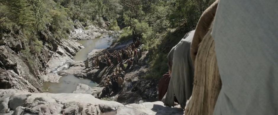 Refugees head down the hill by the waterfall while a larger group waits down below.