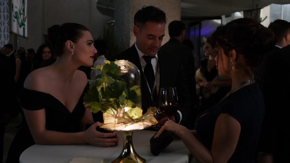 Lena leans against the table where Edge is talking to some woman.