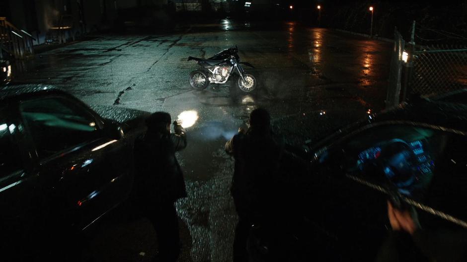 Oliver hides behind his motorcycle as the goons fire at him.