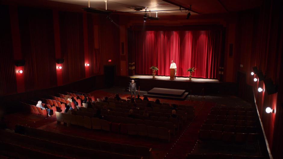 Angus talks to the assembled zombies while in front of the stage where the priest is standing.