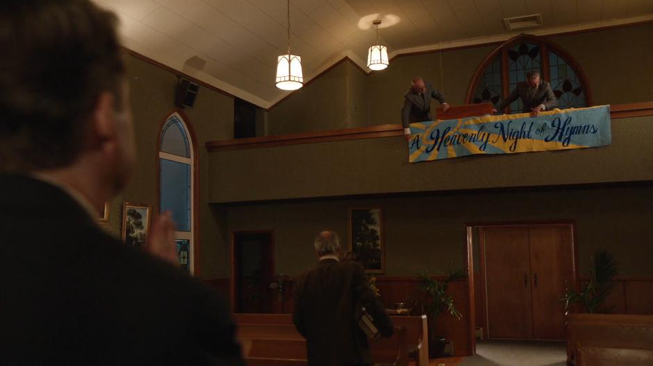 Uncle Lucious watches as two people hang a banner for "A Heavenly Night of Hymns" from the balcony.