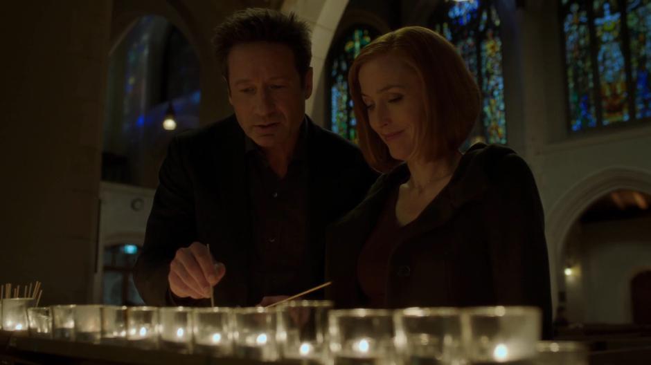 Scully smiles as Mulder attempts to relight her prayer candle.