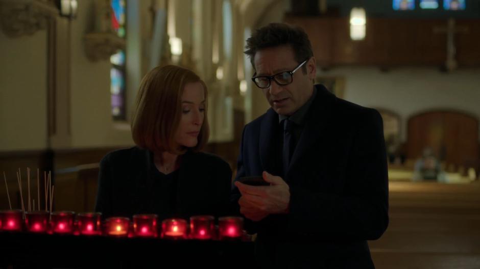 Scully glances over as Mulder looks something up on his phone while wearing his new glasses.