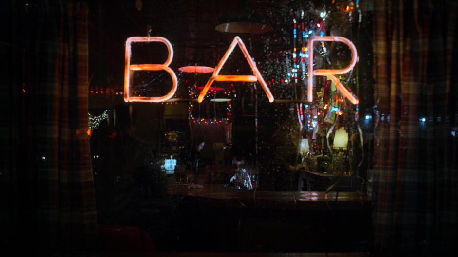 Jessica is visible sitting at the bar through the rain-covered window.