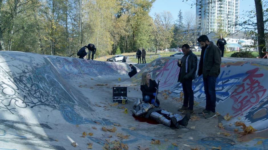 Liv kneels over the body and prepares to remove the sword while Clive and Ravi watch.