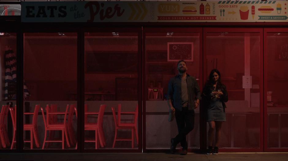 Shaun and Nina lean up agains the snack stand drinking and smoking.