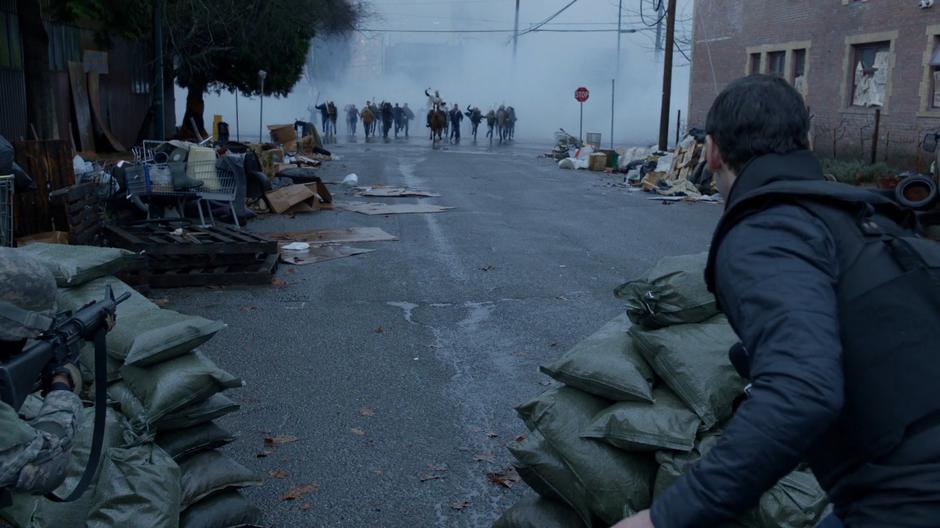 The reporter looks down the street from behind the sandbag line and sees Angus and his congregation emerging from the smoke.