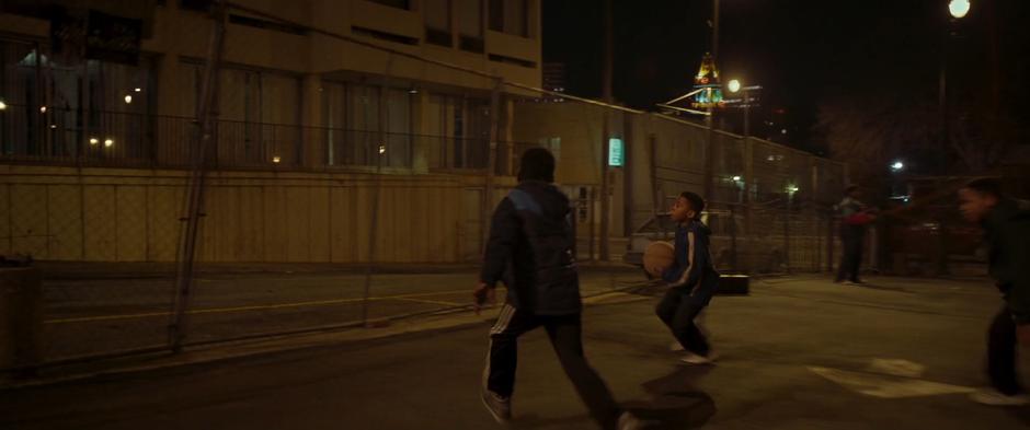 Young Killmonger and his friends play basketball in the lot next to his apartment building.